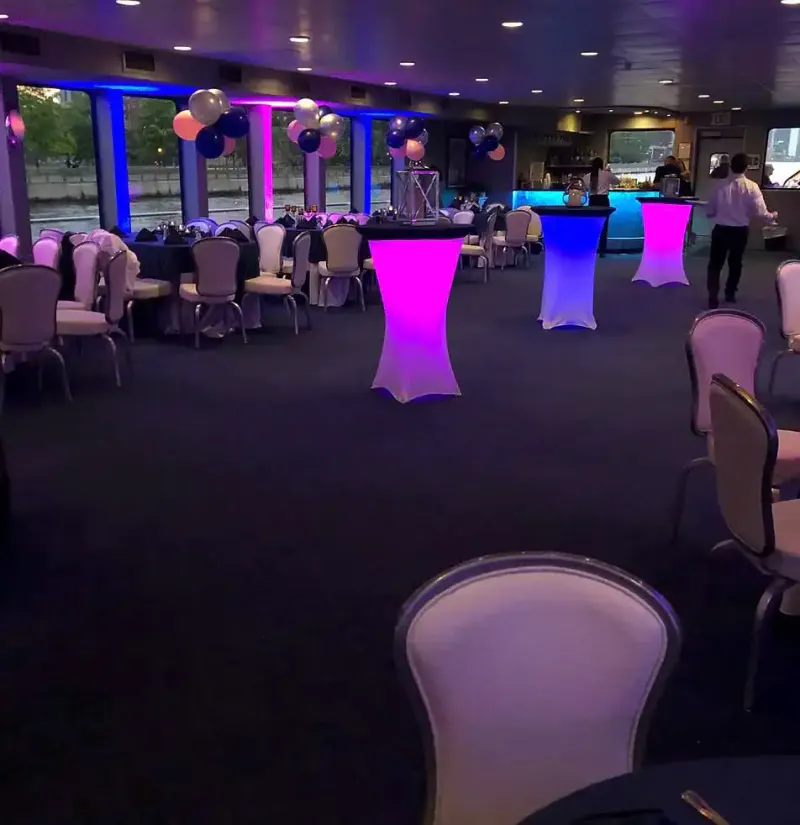 Yacht interior with glowing pink and blue cocktail tables, balloon arches, dining tables and a lit bar by the windows