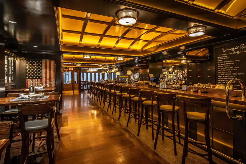Long wooden bar with stools at The Independent NYC, warm gold coffered ceiling, chalkboard beer list and American flag