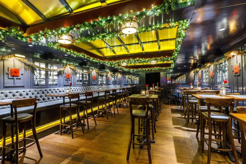 Dining room at The Independent NYC with tufted banquettes, high-top tables and holiday garland lit by yellow ceiling panels
