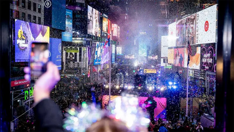 Times Square crowd below glowing billboards with confetti falling on New Year's Eve, a phone raised to film