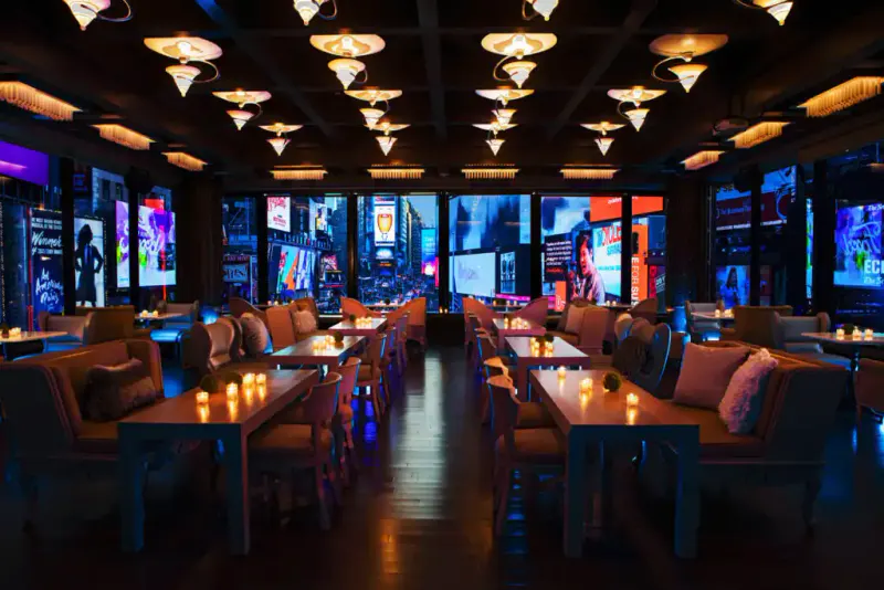 Candlelit dining room with long tables and banquettes facing windows overlooking Times Square billboards at night