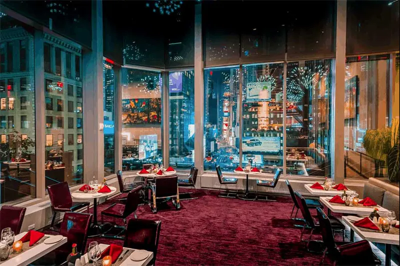 Floor-to-ceiling windows overlooking Times Square lights above a dining room with red carpet and set tables