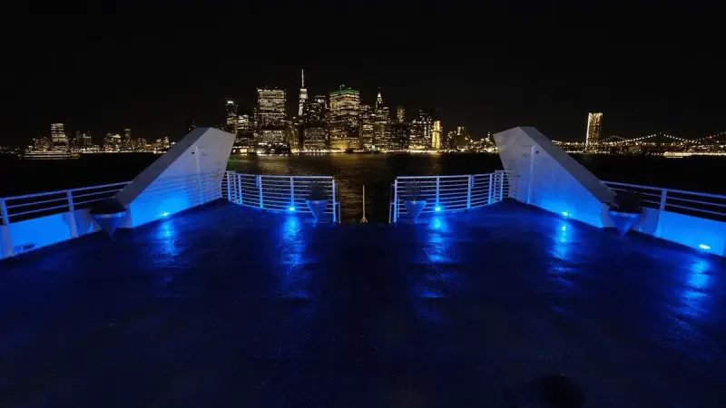 Empty yacht deck lit in blue at night with the lower Manhattan skyline glowing across the water