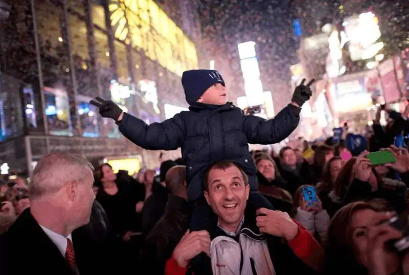Child on a father's shoulders raising arms amid confetti in a Times Square New Year's Eve crowd