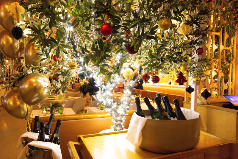 Champagne bottles chilling in buckets under holiday trees with ornaments and string lights in a golden dining room