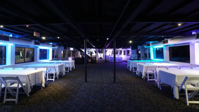 Interior deck of a party cruise yacht with white-draped tables, folding chairs and blue LED lighting along the windows