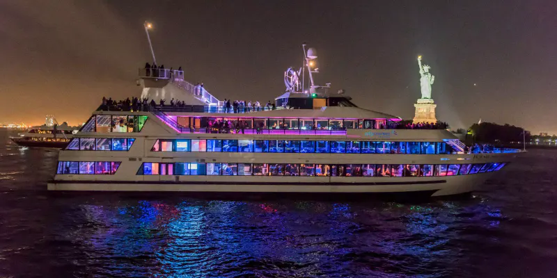 Hornblower Infinity yacht lit up in blue and purple at night with guests on deck and the Statue of Liberty behind