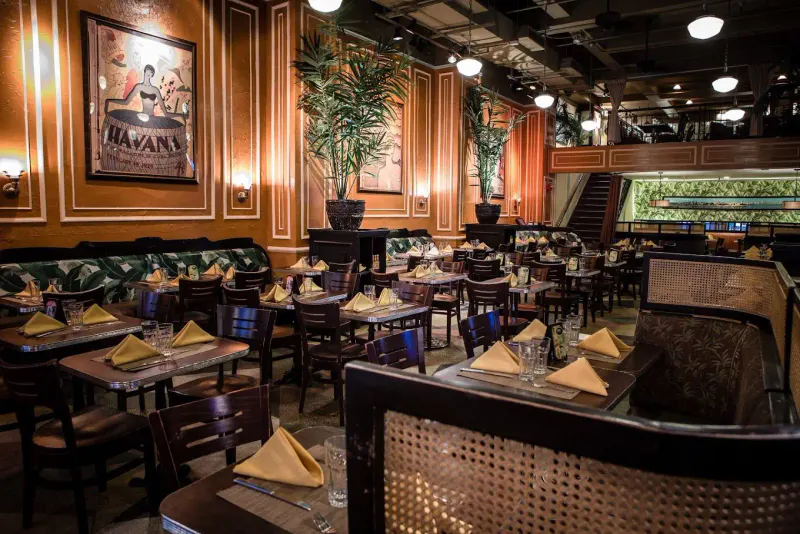 Havana Central dining room with set tables, yellow napkins, palm plants and a vintage Havana poster on warm orange walls