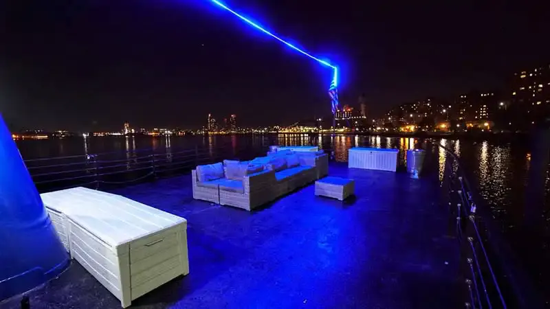 Open top deck of a party cruise at night with lounge seating, blue lighting and city skyline across the water