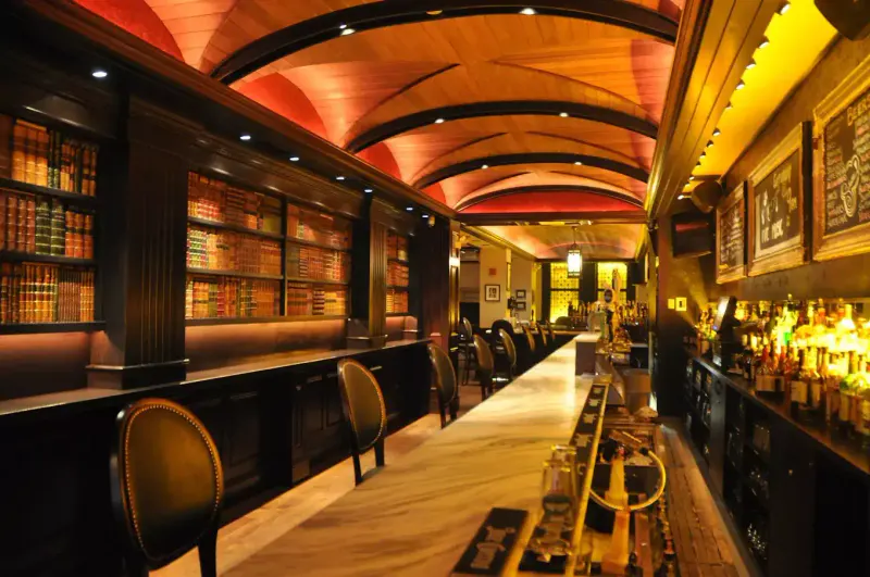 Long marble bar at The Long Room NYC with vaulted wood ceiling, bookshelves and leather bar stools