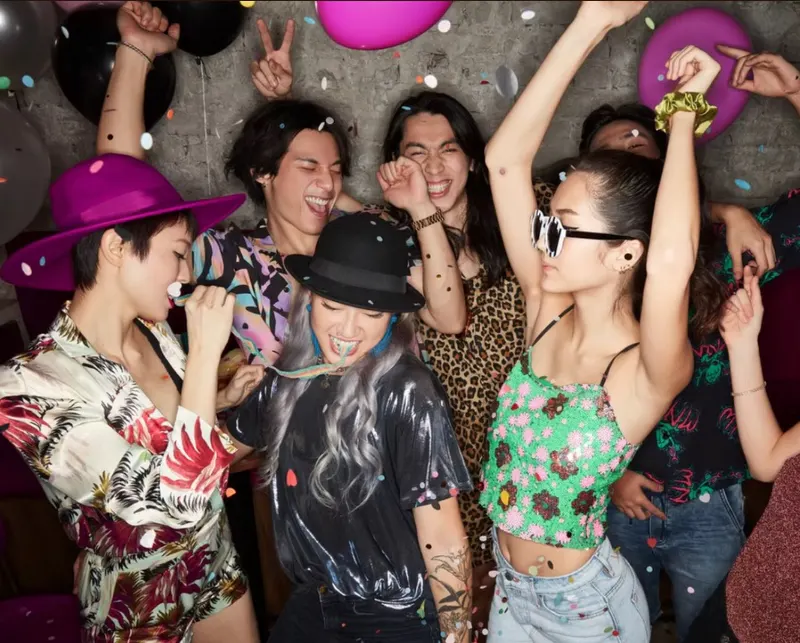 Group of friends dancing under falling confetti with balloons, party hats and sunglasses at a nightclub party