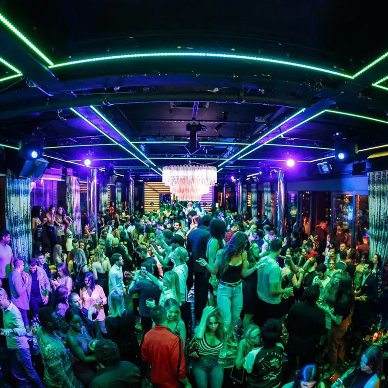 Packed nightclub dance floor under green and purple neon ceiling lights and a crystal chandelier