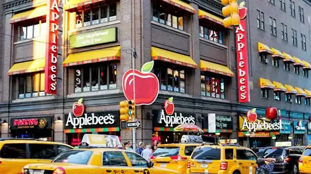 Exterior of Applebee's in Times Square with large red apple signs, yellow awnings and taxis passing below