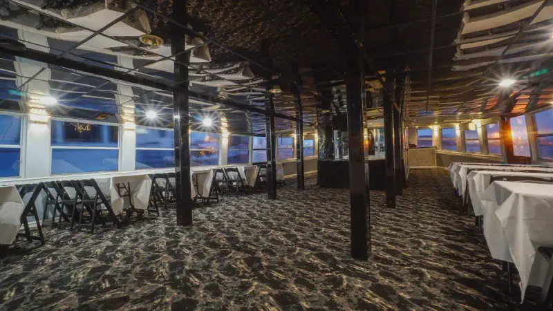 Interior deck of the Lucille Yacht with white-draped dining tables, folding chairs and windows at dusk