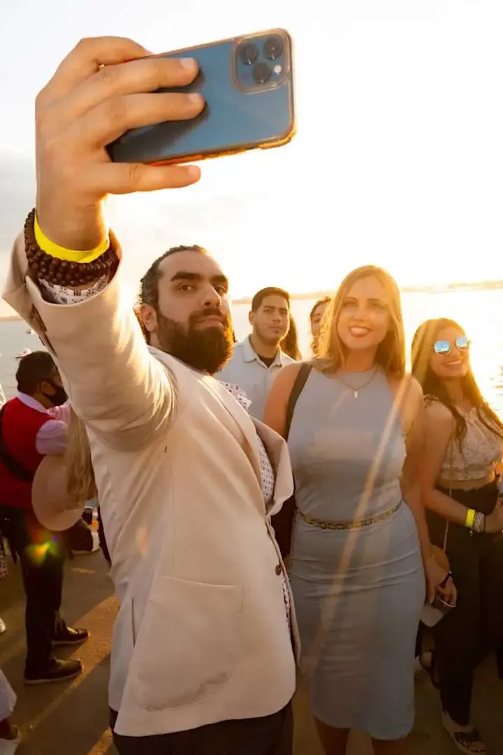 Guests taking a group selfie on the deck of a party cruise at golden hour with water behind them