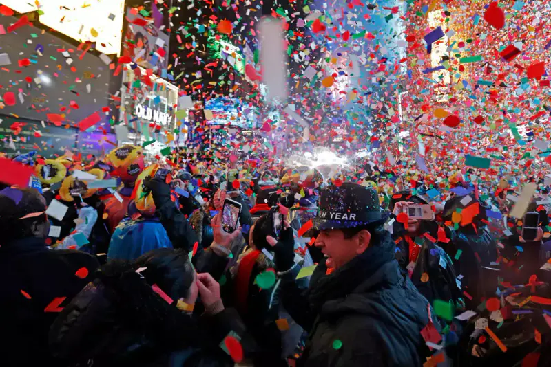 Crowd in Times Square at midnight under falling confetti, one reveler in a New Year hat raising a phone