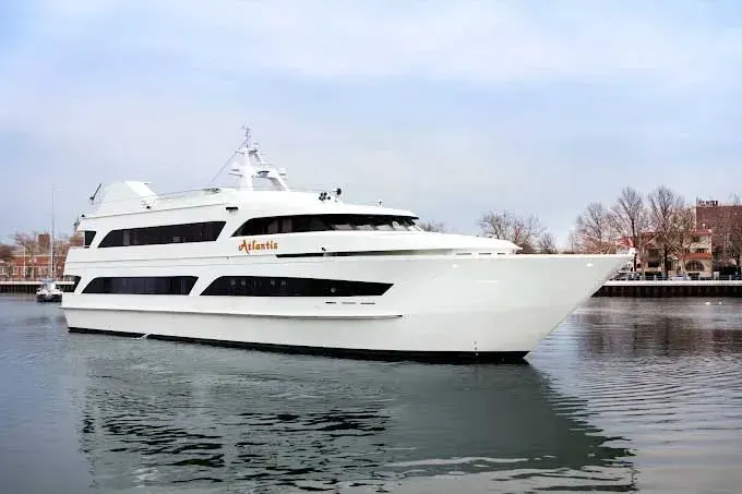 White multi-deck Atlantis yacht docked on calm water with bare trees and buildings along the shore