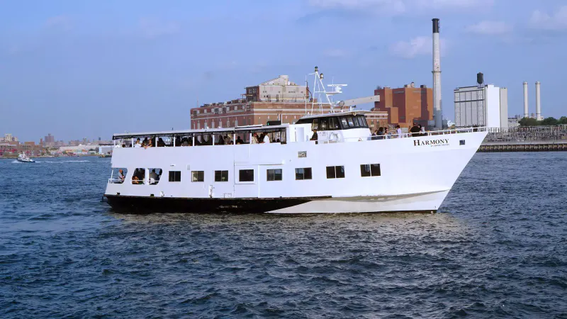 Harmony yacht cruising NYC waters with guests on the open upper deck and a waterfront skyline behind