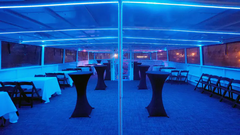 Enclosed yacht deck with blue LED lighting, black cocktail tables and folding chairs along the windows