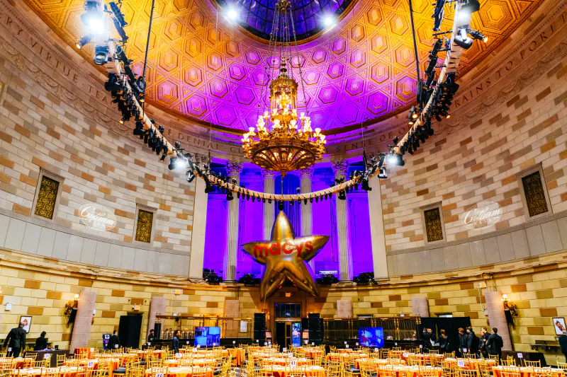 Gotham Hall grand ballroom with purple-lit domed ceiling, chandelier, gold star balloon and round banquet tables set for