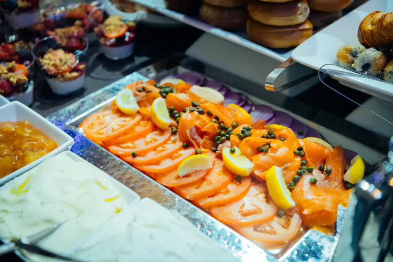 Brunch buffet platter of smoked salmon with sliced tomato, lemon wedges and capers beside bagels and pastries