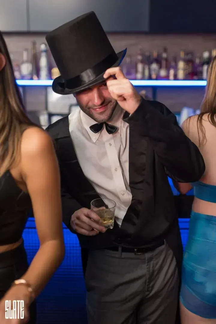 Man in top hat and bow tie tipping his hat with a cocktail at the bar during a Halloween party at Slate NY