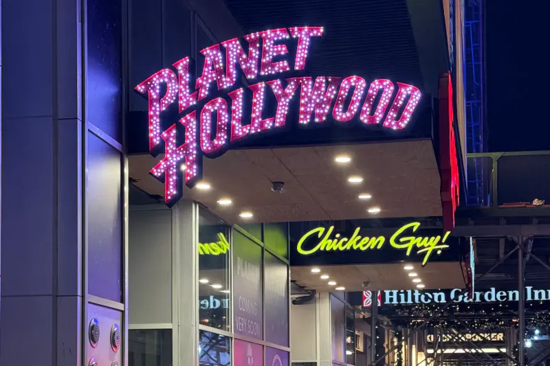 Lit-up Planet Hollywood marquee and neon Chicken Guy sign above the entrance in Times Square at night