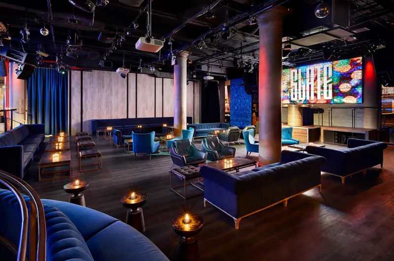 Interior of Slate NY with blue velvet sofas, candlelit tables and a glowing video wall above the stage
