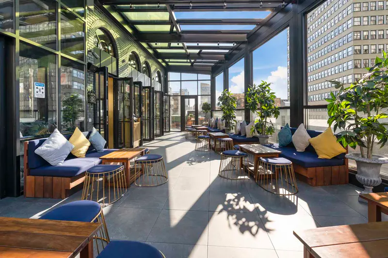 Sunlit glass-enclosed rooftop lounge at Elsie Rooftop with navy banquettes, wood tables and city views