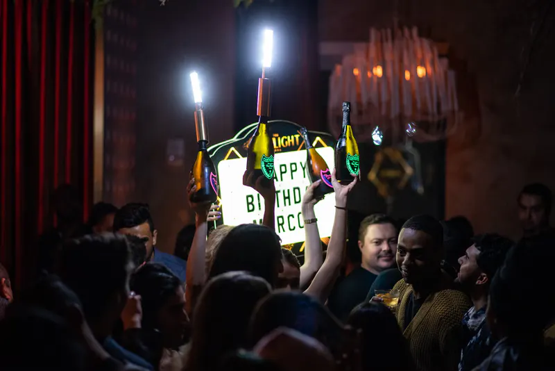 Bottle service sparklers and a lit happy birthday sign carried through a crowded nightclub floor