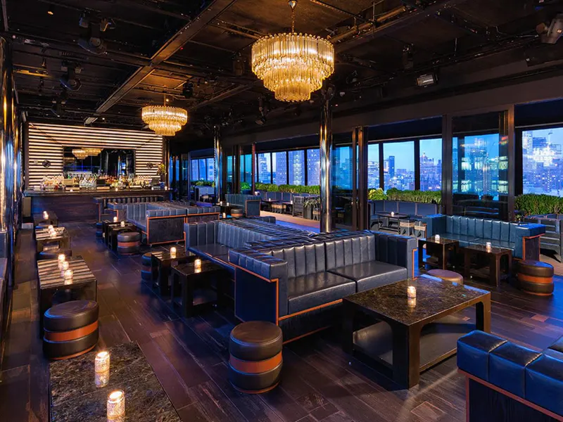 Rooftop lounge interior with crystal chandeliers, blue leather banquettes and windows overlooking the NYC skyline