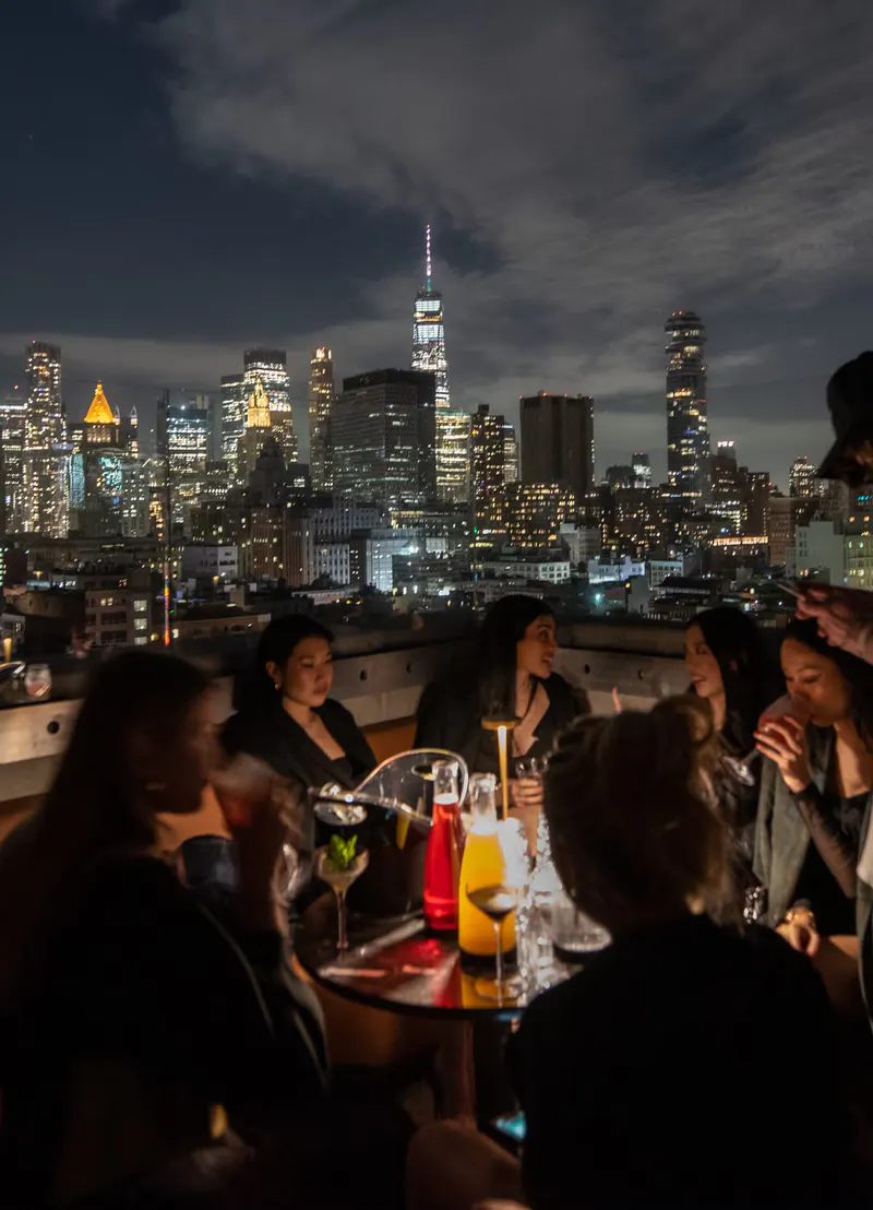 Guests with cocktails at a rooftop table overlooking the lit downtown Manhattan skyline at night